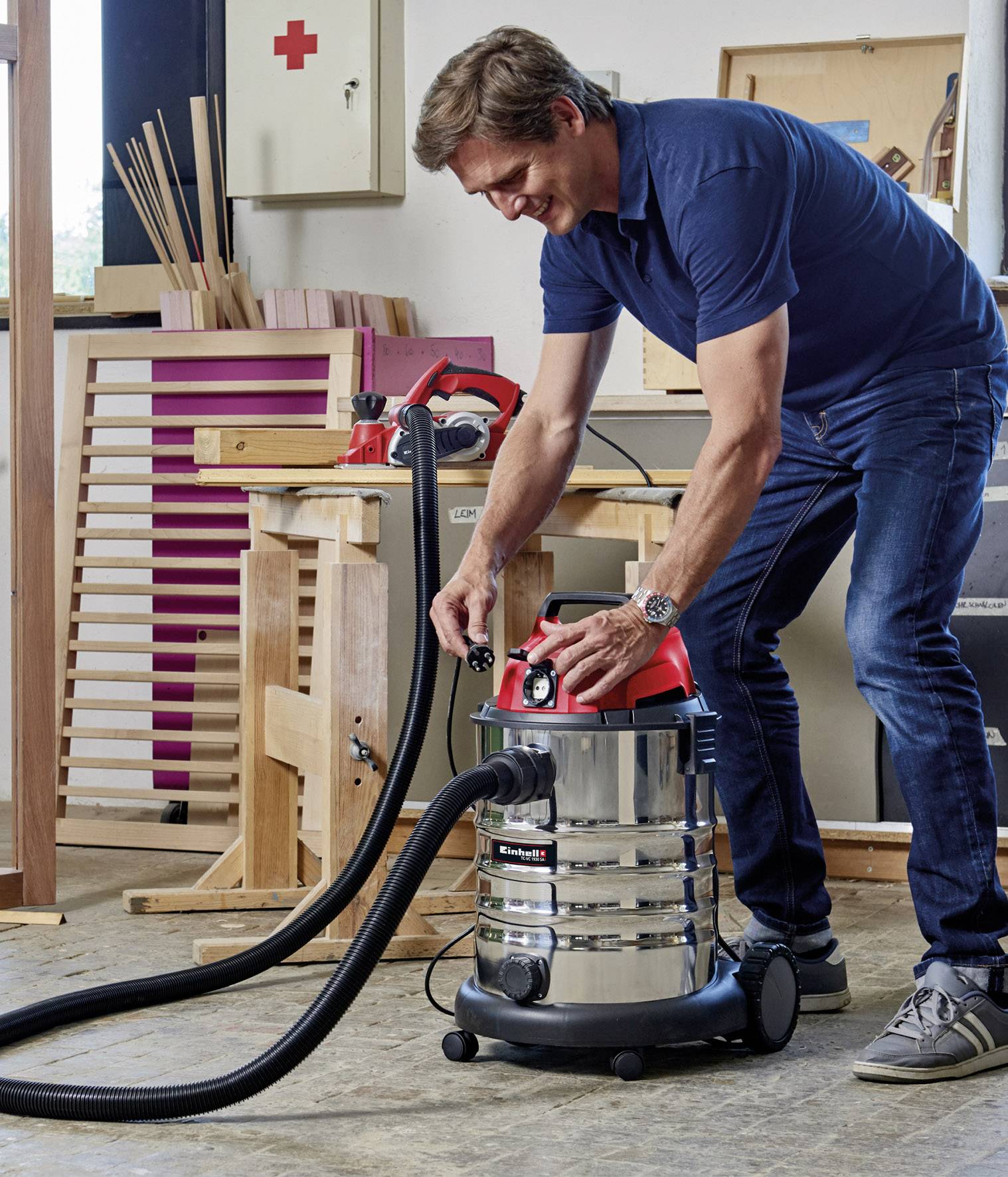 A man in a workshop is operating an industrial vacuum cleaner with a red lid, surrounded by wooden materials and tools.