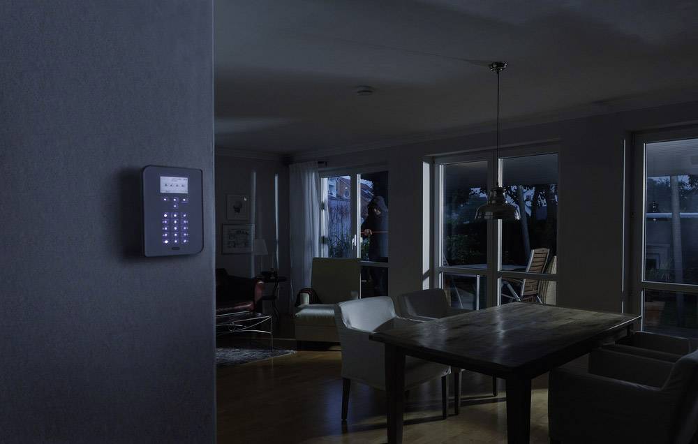 A burglar stands by the window of a dark living room at night. An illuminated security control panel is visible on the wall in the foreground.
