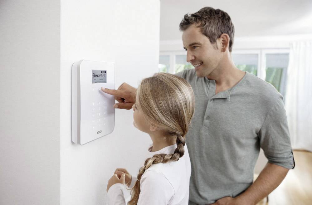 A man and a girl are standing in front of a home alarm system. The man is showing the girl how to operate the numbers on the keypad.