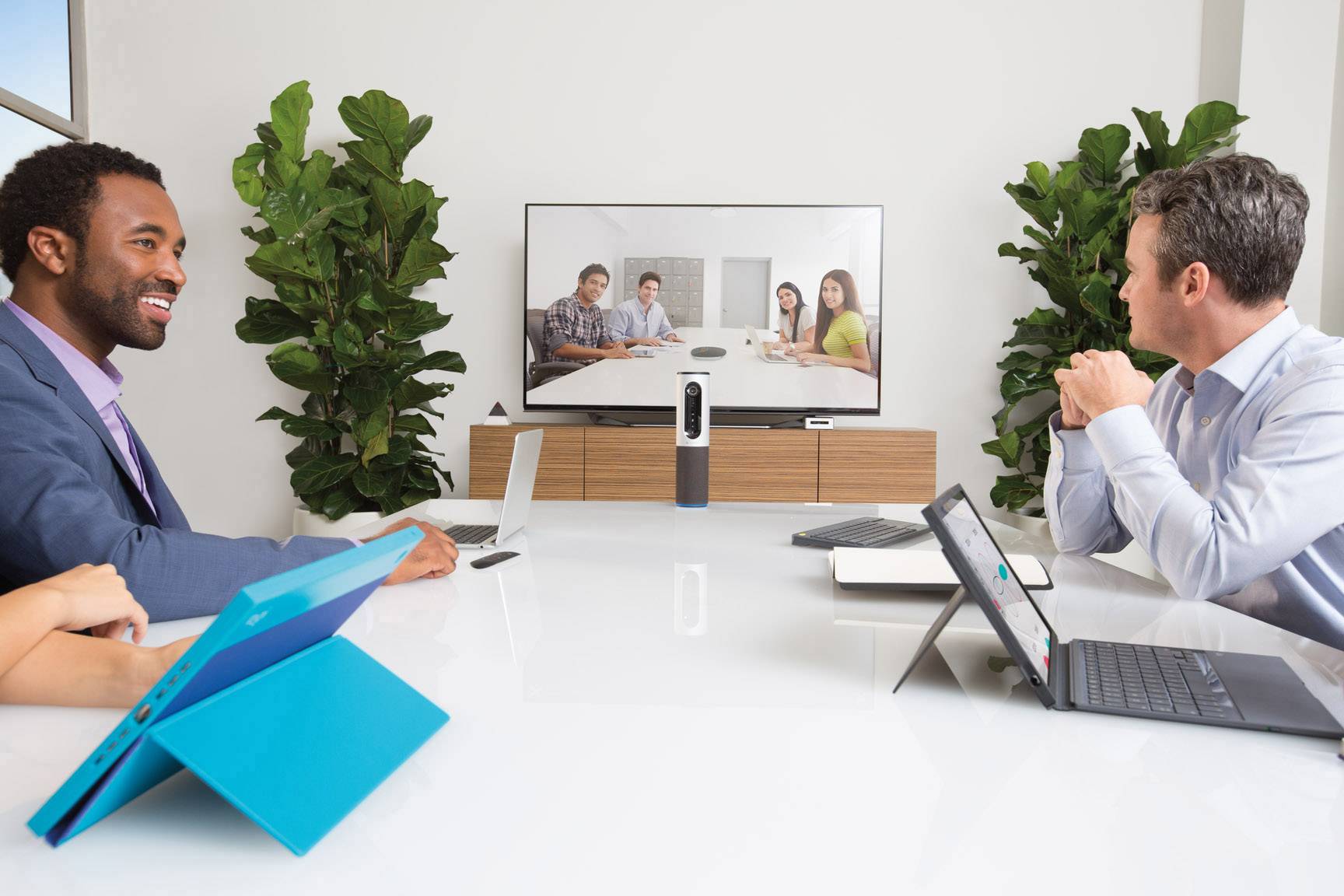 People in a meeting room are conducting a video conference. Two visible participants are working on laptops, while additional participants are displayed on the screen.