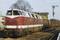 A red diesel locomotive is pulling a goods train through a railway station. Trees and a signal can be seen in the background.