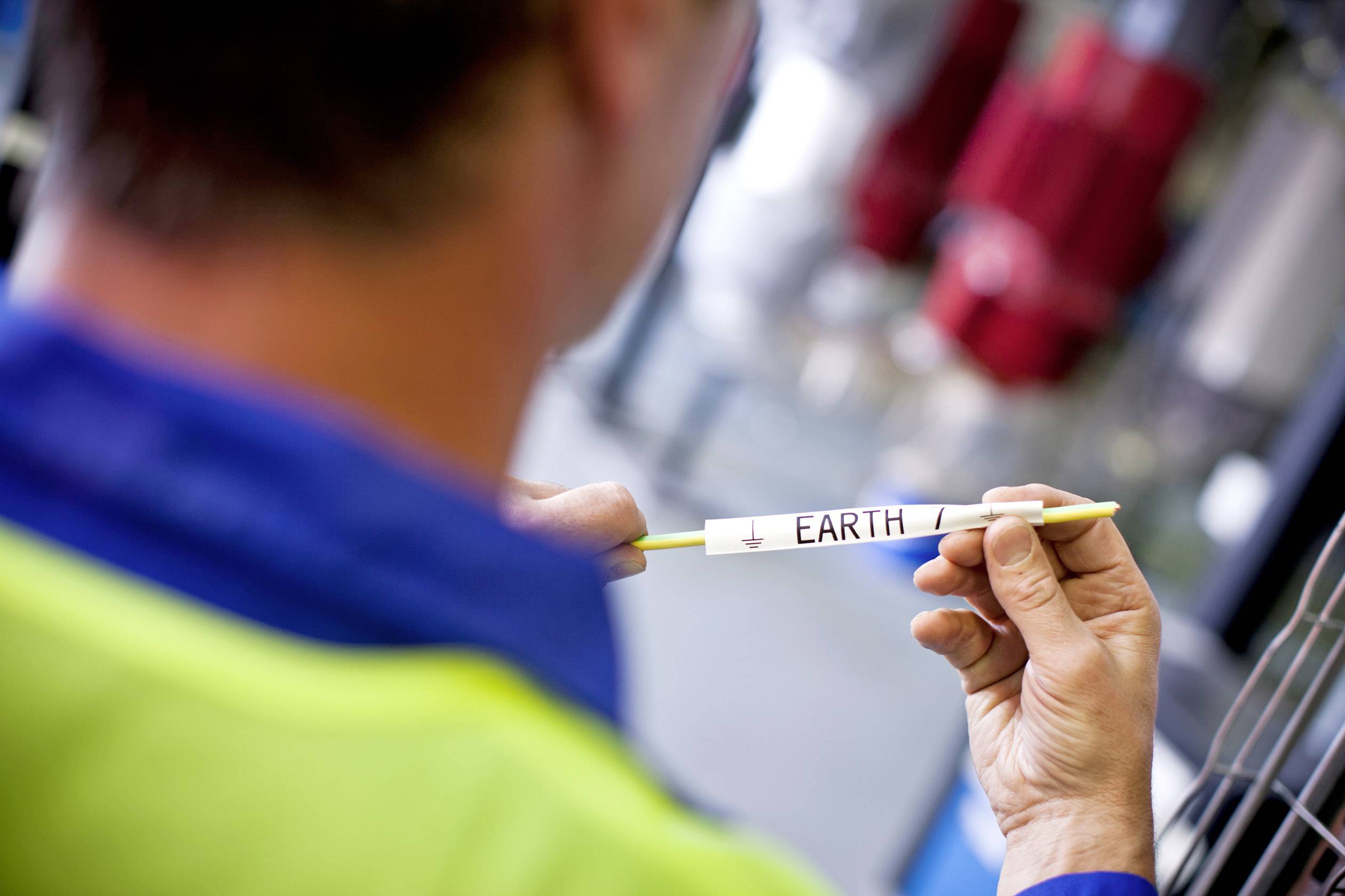 A man is holding a yellow pen with the inscription 'EARTH'. He is wearing a work jacket, with blurred tools in the background.