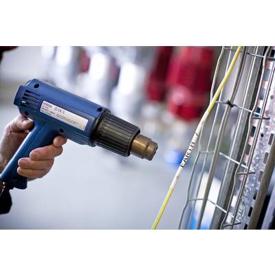 A person is using a blue heat gun to seal heat-shrink tubing on cables. Blurred objects are visible in the background.