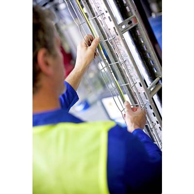 A man in workwear is measuring a metal grid with a tape measure. Industrial elements are blurred in the background.