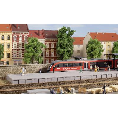 A model railway scene depicts a red train at a platform in a city with historic buildings and trees.