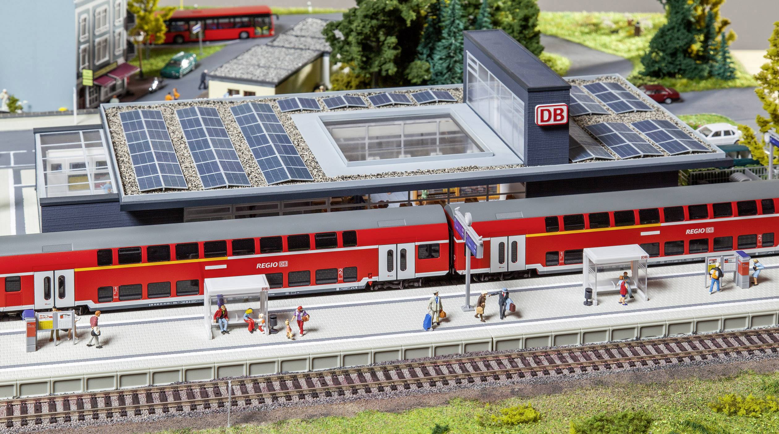 A model railway station with red trains and a platform. Buildings with solar panels. People waiting on the platform.