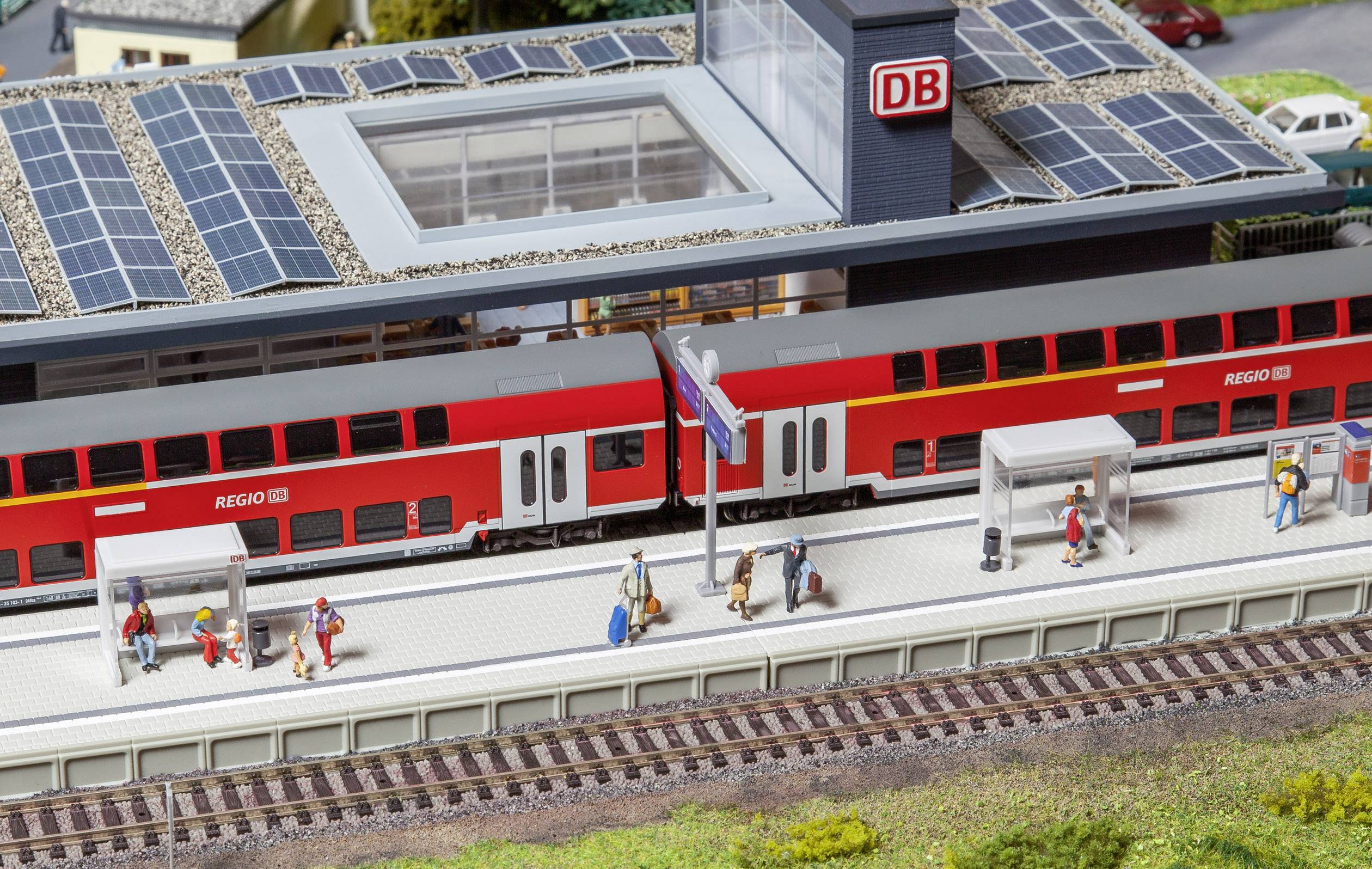 A red double-decker train at the railway station, people waiting on the platform. Buildings with solar panels in the background.
