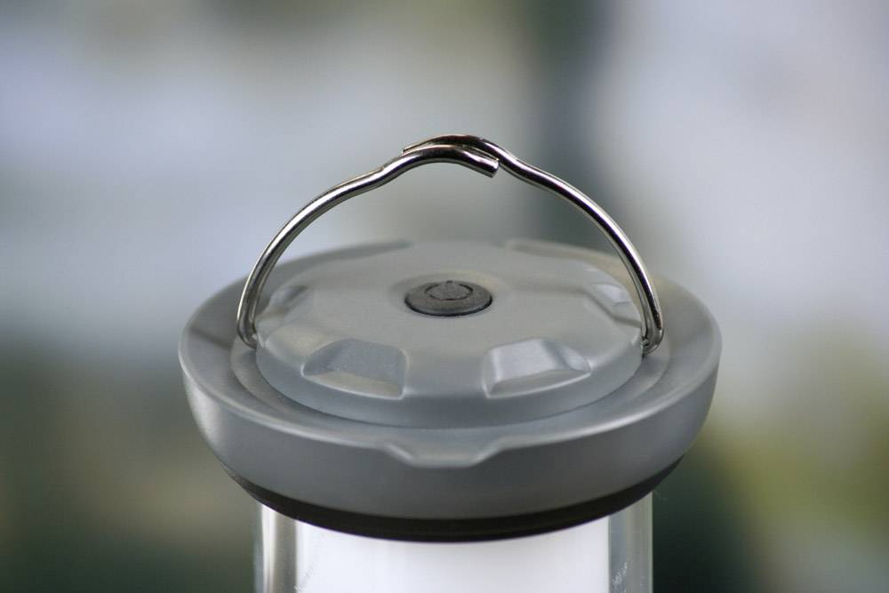 A grey lantern with a folding handle and a visible on/off button on the top, against a blurred background.