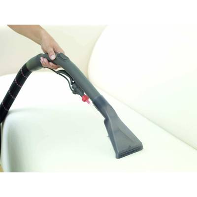 A person is using a steam cleaner to clean a light-coloured sofa. The steam cleaner has a wide black attachment.