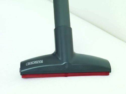 A vacuum cleaner attachment is sucking a white surface. The attachment is dark with a red rubber strip and the 'Thomas' brand.