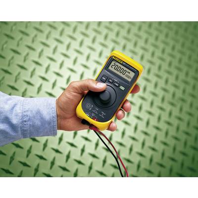 A hand is holding a yellow digital multimeter displaying '20000 mA' against a green, textured background.