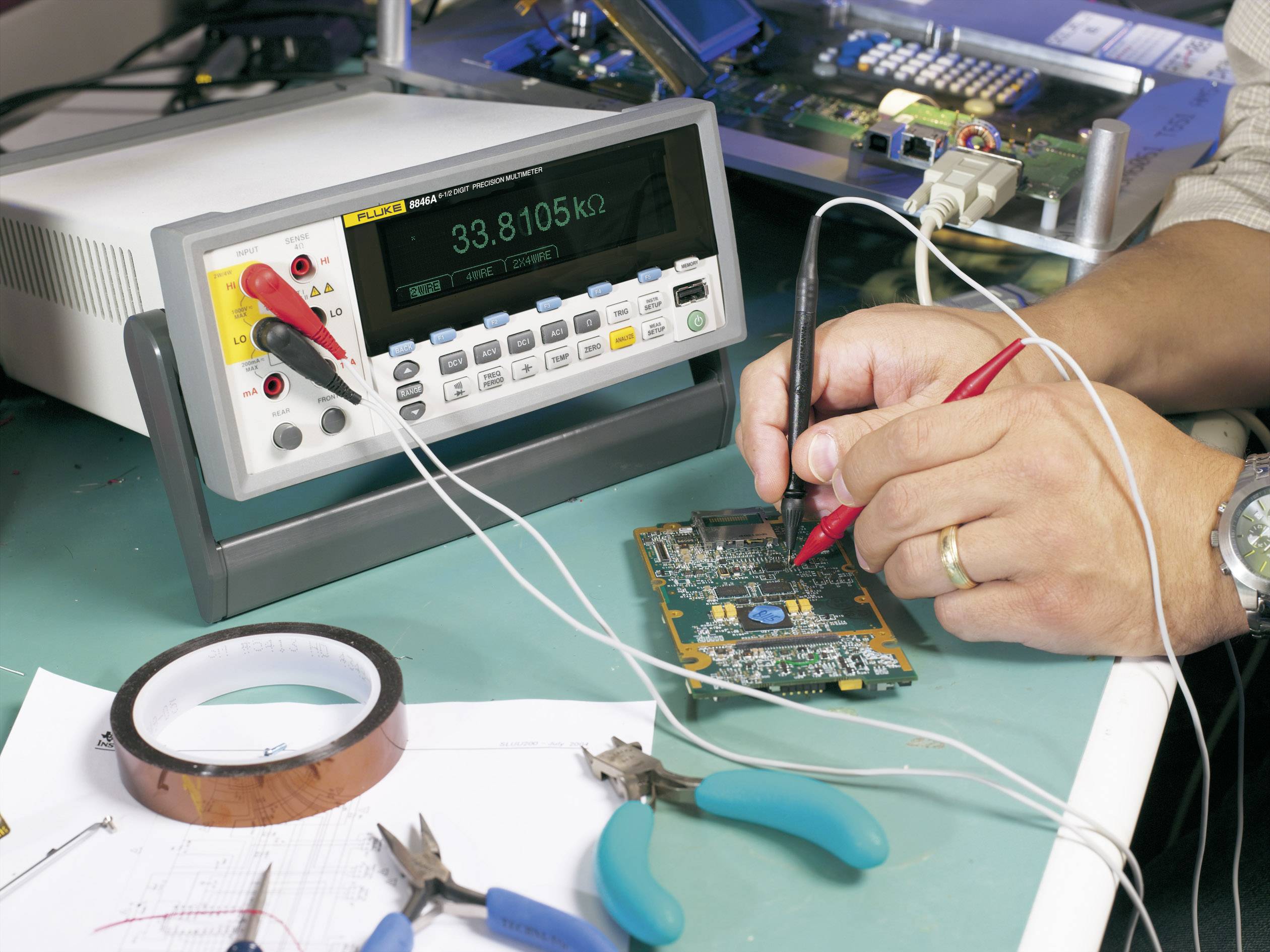 A technician is measuring the electrical voltage on a circuit board using a multimetre. Tools and circuit details are visible in the background.