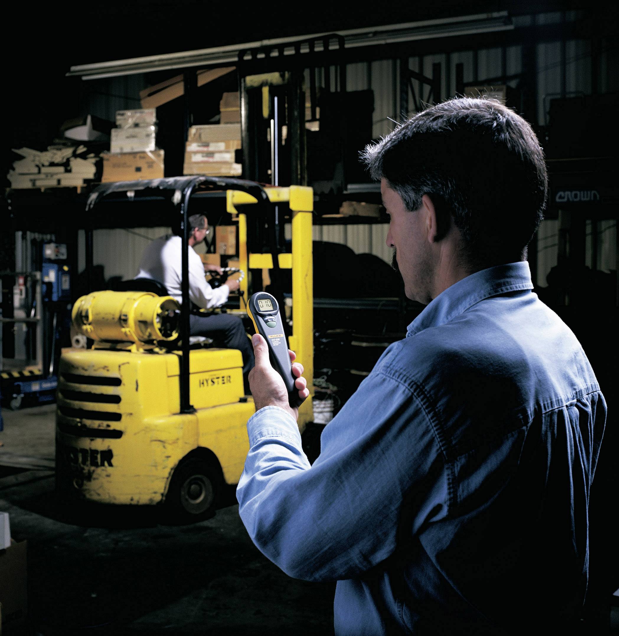 A man is using a smartphone in a warehouse while a forklift driver is working. A forklift is visible in the background, with shelves stacked with cardboard boxes.