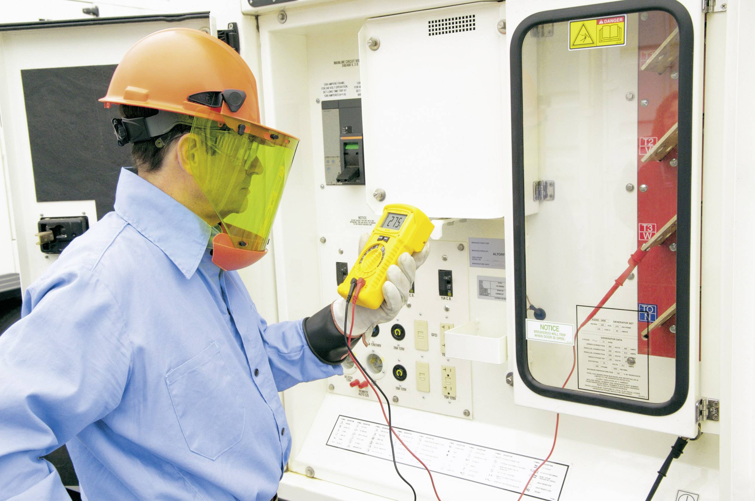 A worker is wearing protective clothing and measuring electrical voltage at a control cabinet using a multimetre.