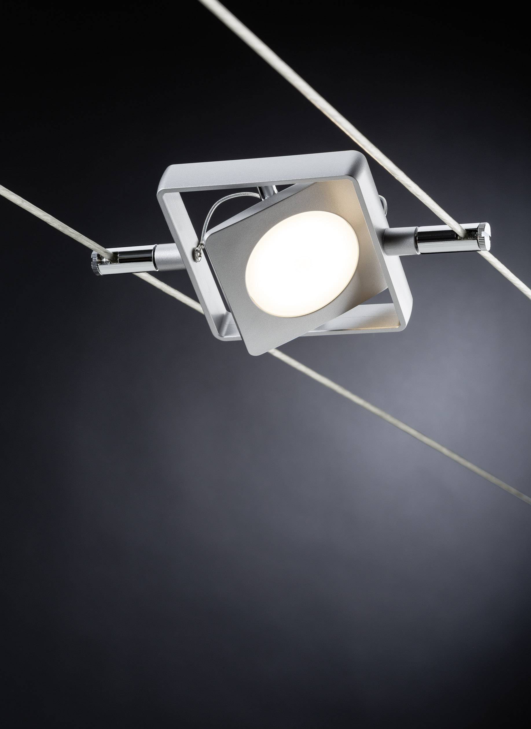 A modern ceiling spotlight in a square housing hangs from two thin cables against a dark background.
