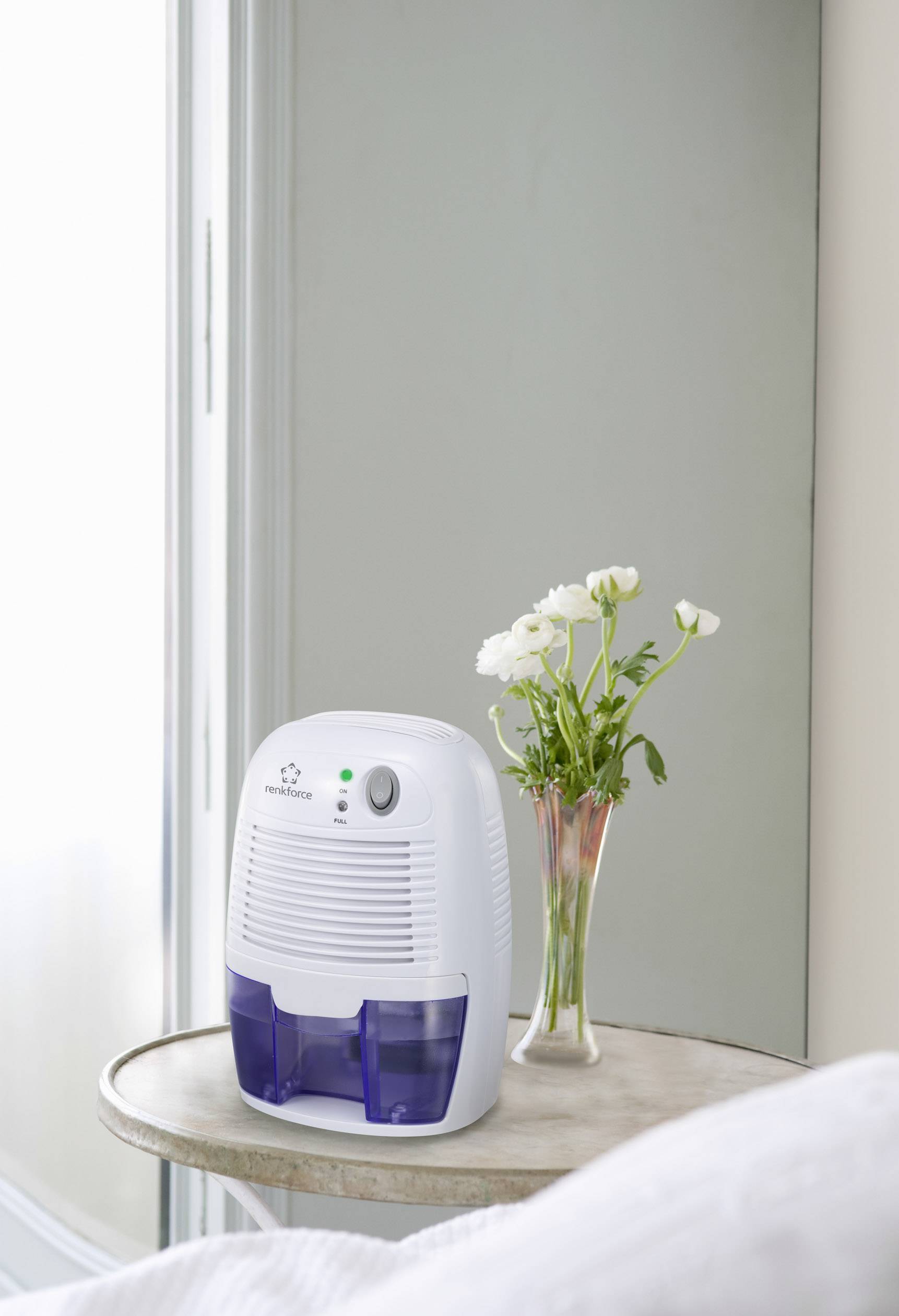 A dehumidifier stands on a table beside a window, with a vase of white flowers next to it.