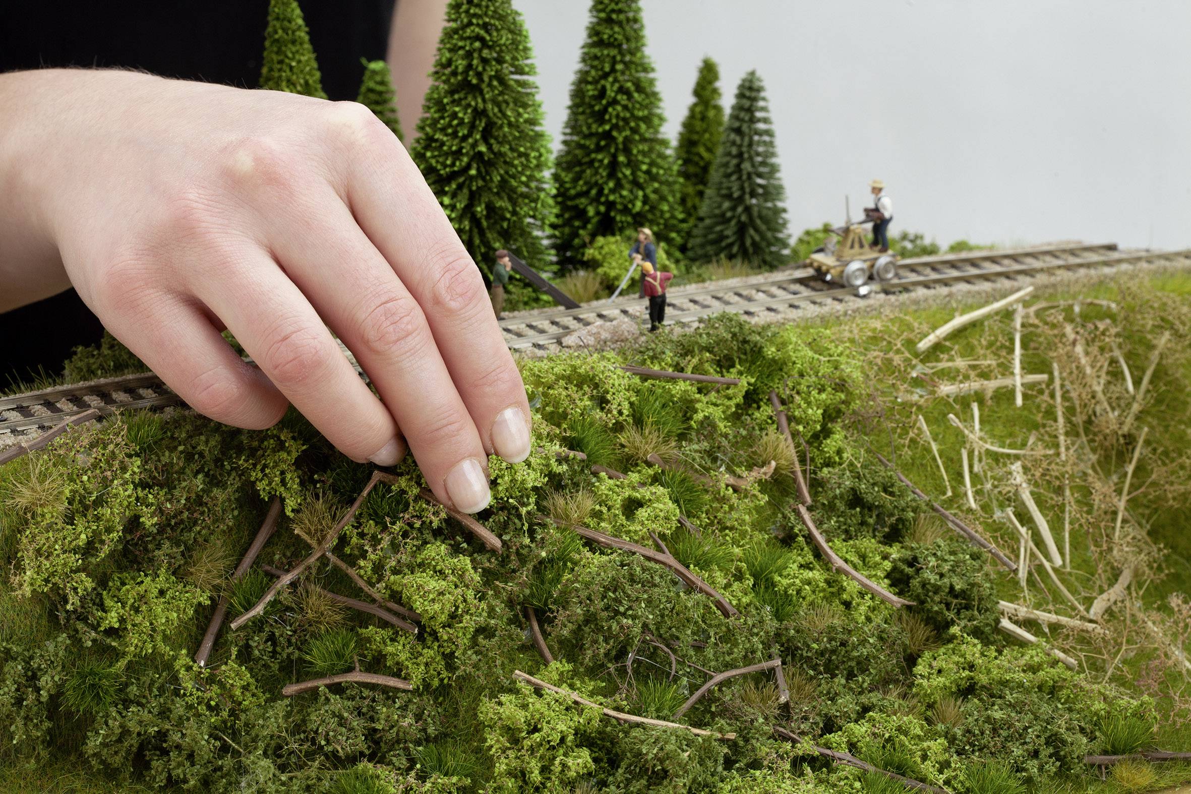 A hand arranges miniature trees on a modelled hill beside a model railway track with small figures.