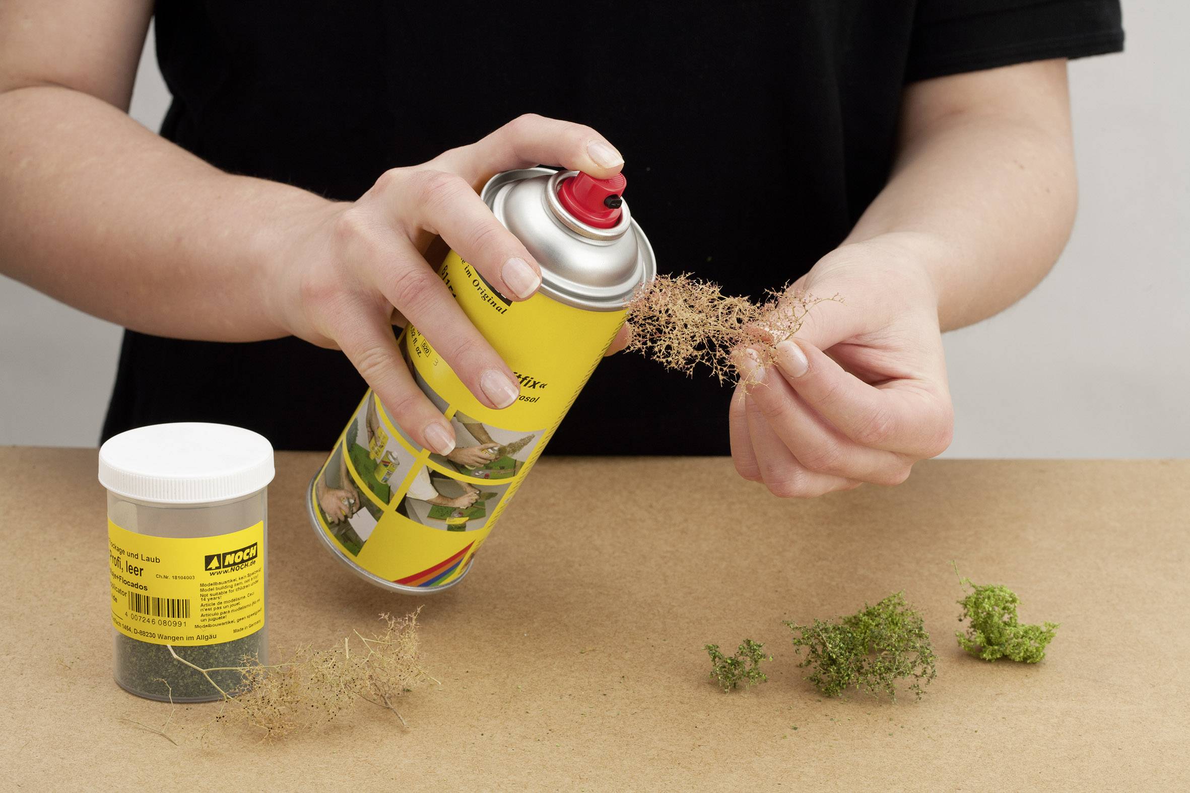 A person is spraying yellow paint from a spray can onto model tree moss. An open box of model tree moss sits next to it on a table.
