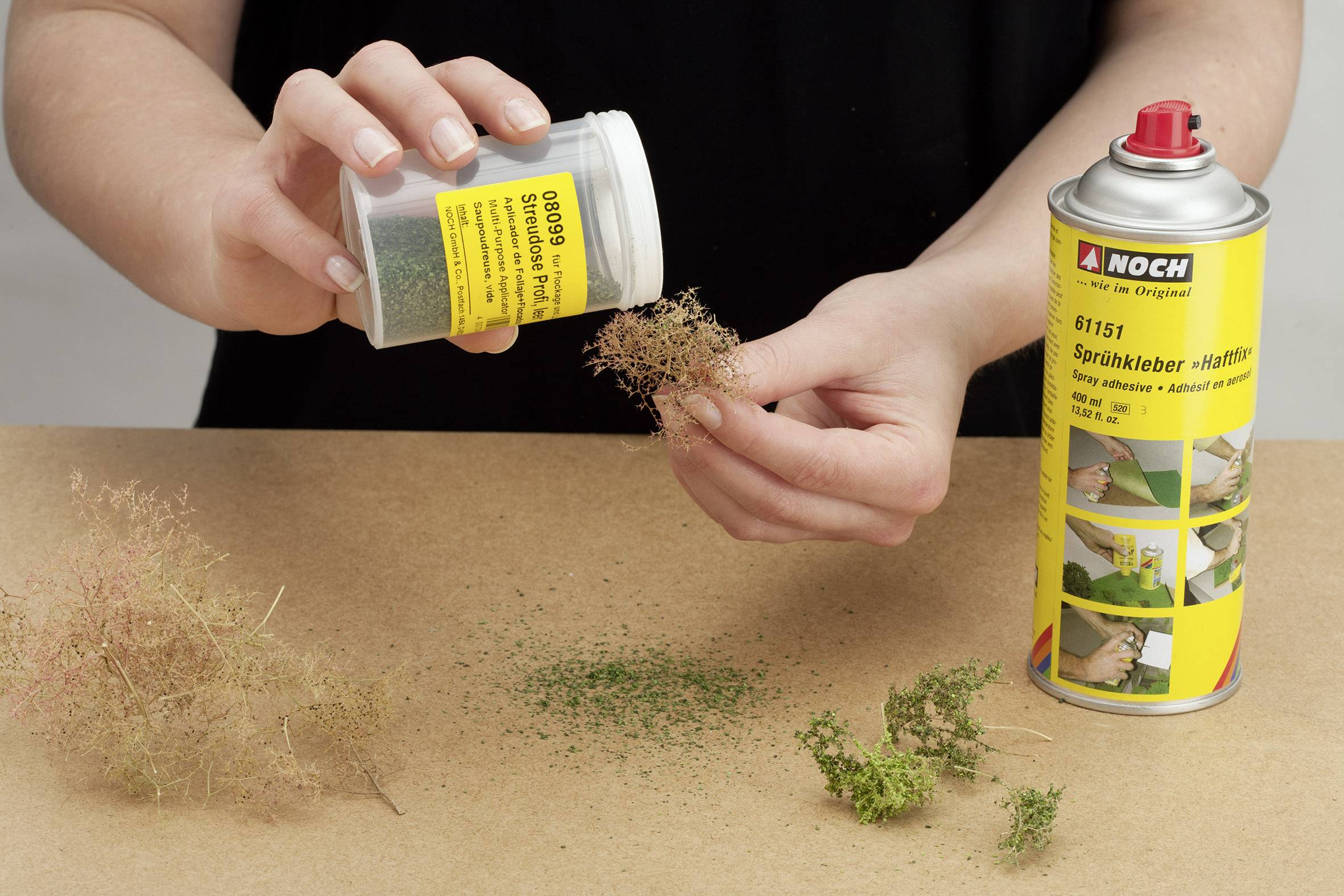 A person is sprinkling green powder onto moss, with a spray adhesive can beside them.