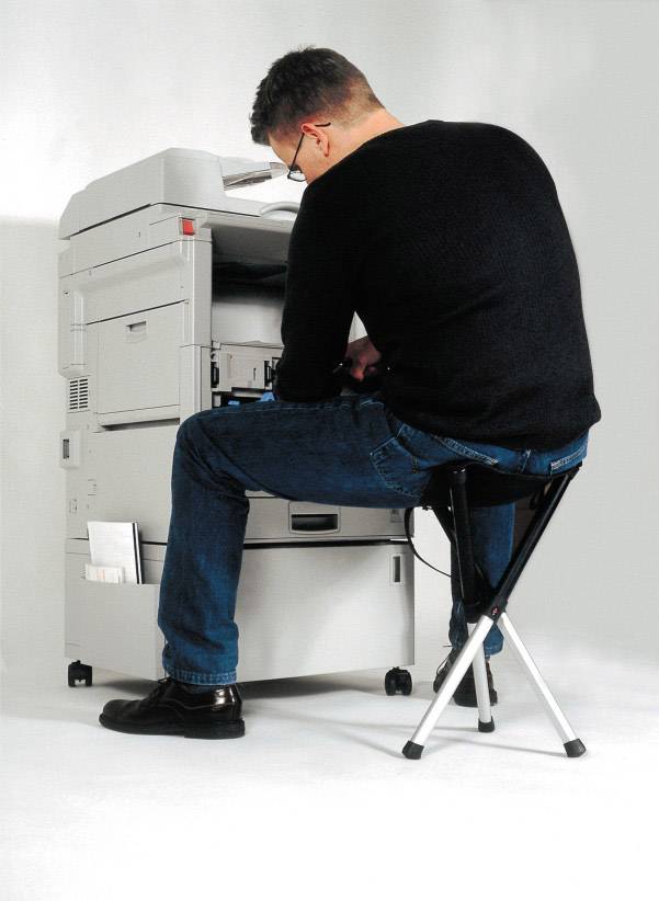 A man is sitting on a stool and operating a photocopier.