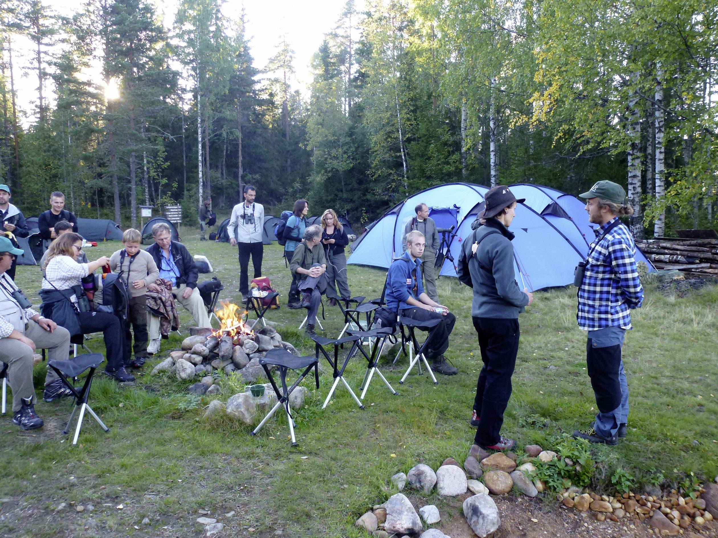 A group of people are sitting around a campfire outdoors. Blue tents stand in the background. Two individuals are standing and chatting with each other.
