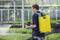 A person is spraying plants in a greenhouse with a yellow backpack sprayer, surrounded by green vegetation.