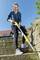 A woman is using a yellow garden tool to remove weeds on a stone staircase in front of a house with a red roof.