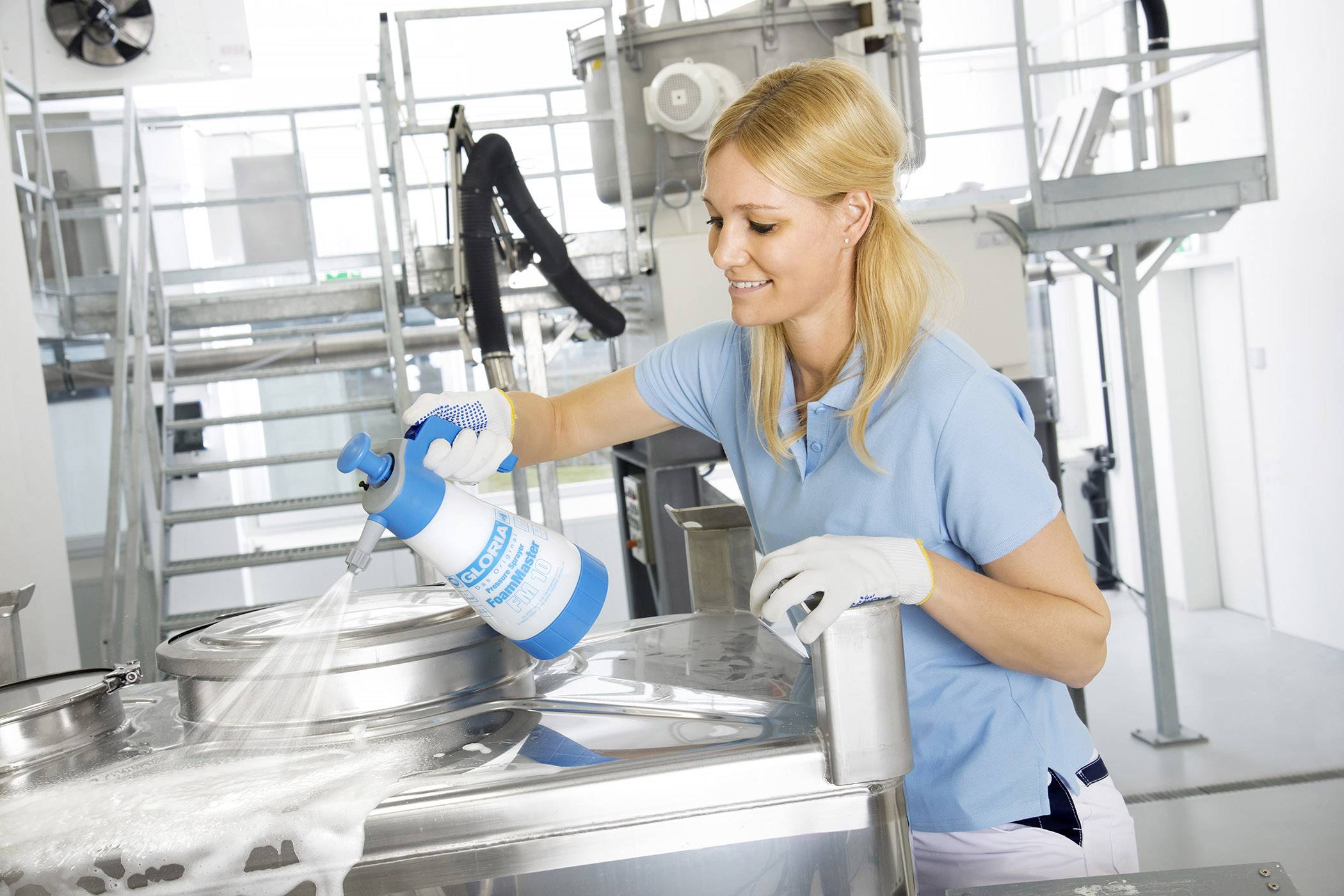 A woman in blue clothing is cleaning a metal machine with a spray cleaner in an industrial environment.