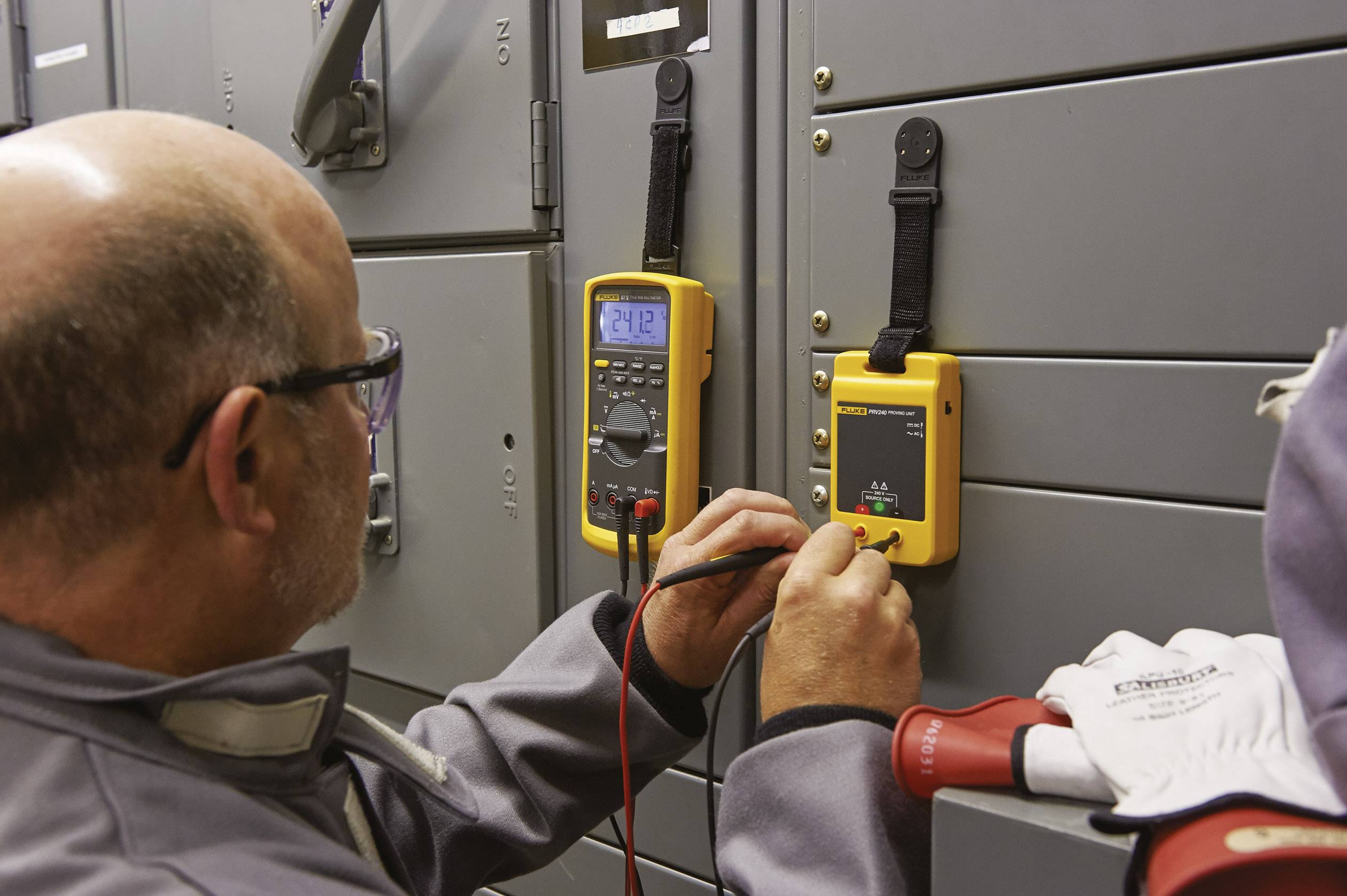 A man is measuring the voltage in an electrical cabinet using a digital multimeter. Safety glasses and gloves ensure his protection.