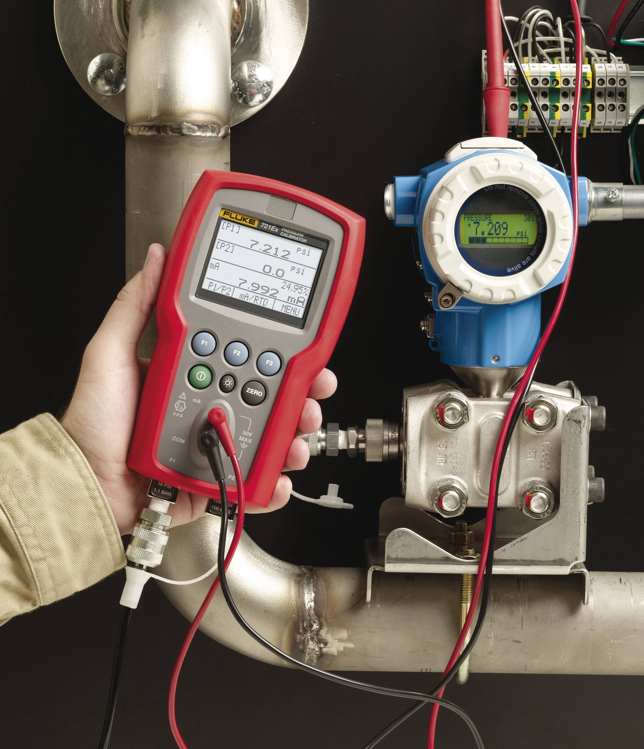 A person is holding a red measuring device connected to a blue device on a pipeline. The device is displaying measurement readings.