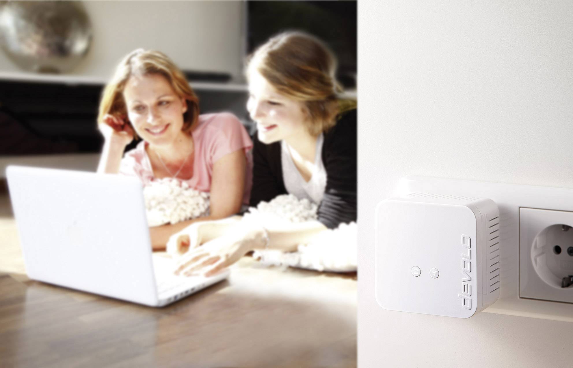 Two people are lying on the floor and sharing a laptop. A Devolo device is plugged into a socket in the foreground.
