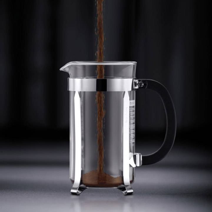 Glass coffee press with a black handle and silver rim, coffee being poured from above, dark background.
