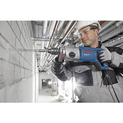 A man is working with a blue Bosch drill hammer on a brick wall in an unfinished building. He is wearing protective clothing and a hard hat.