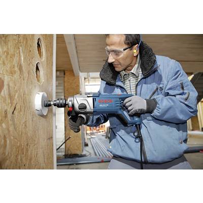 A man in protective clothing is drilling a large hole into a wooden board on a construction site using a blue electric power tool.