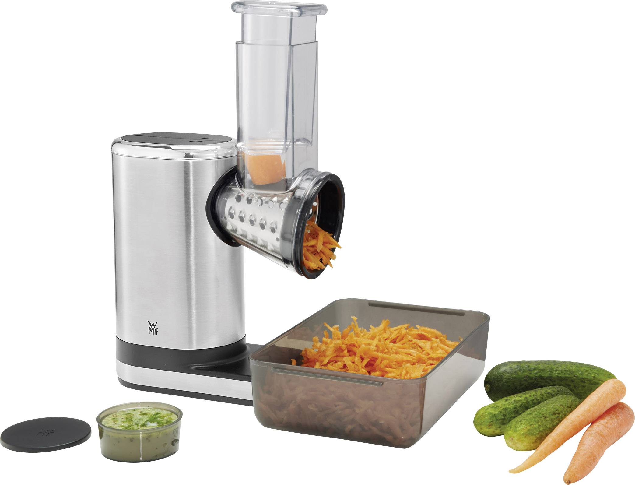 Electric vegetable chopper with stainless steel casing grates carrots into a rectangular dish. Cucumbers and a carrot lie beside it.
