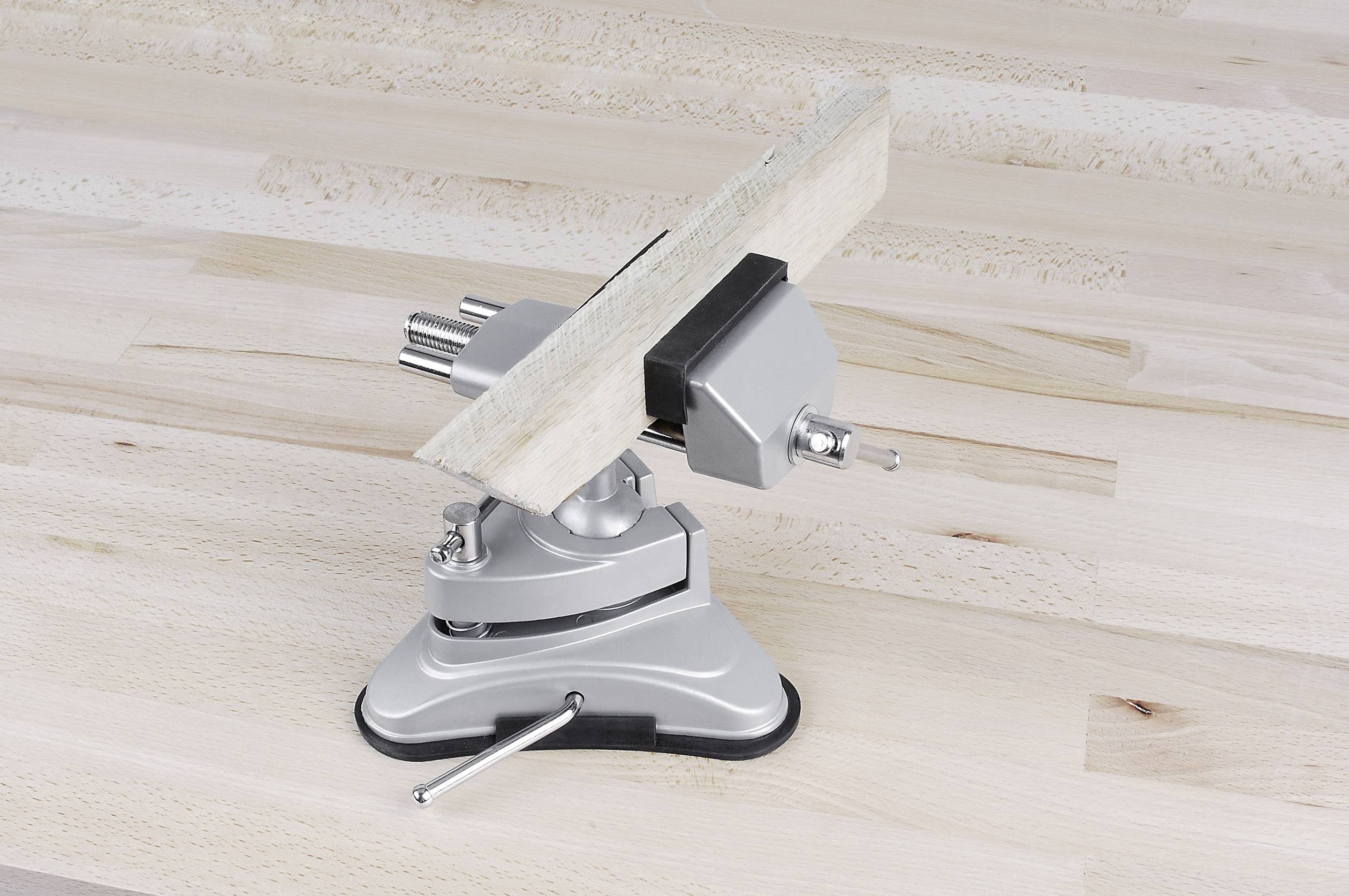 A silver bench vice holds a piece of wood horizontally on a light wooden surface.