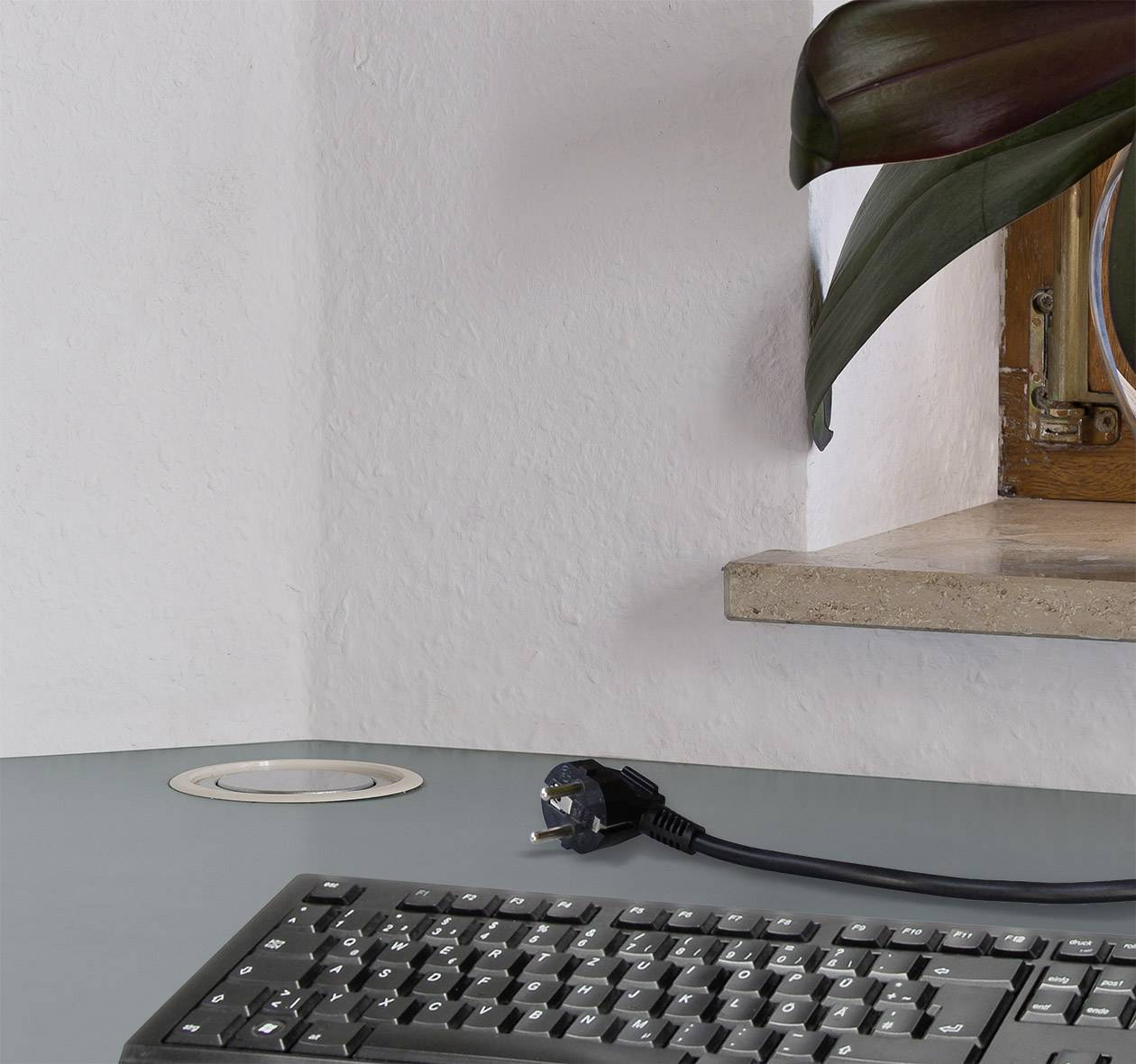 A room with a plant on a wall shelf and a black computer keyboard on a grey table. The power socket is visible.