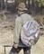 A person in outdoor attire is carrying a large round, camouflaged rucksack and holding a walking stick. Trees in the woods are visible in the background.
