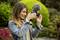 A woman is holding a camera with a microphone in a garden. She is smiling and focusing. Green bushes are visible in the background.