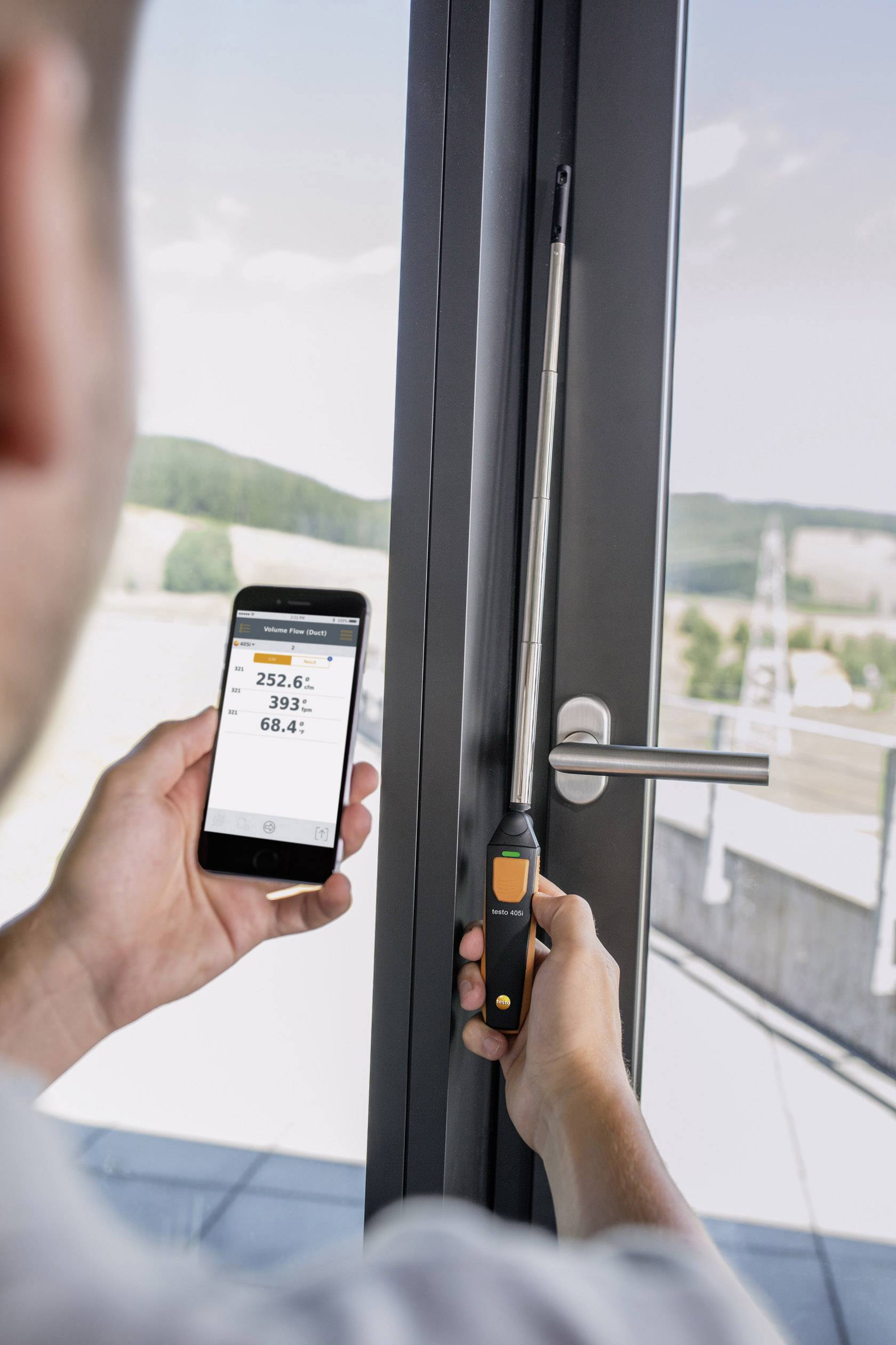 A person is measuring the moisture of a window with a device that transmits the readings to a smartphone. Background: landscape.
