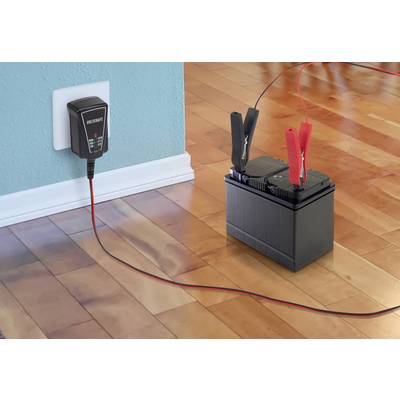 A battery charger is plugged into a wall socket and is charging a car battery via red and black clamps on a wooden floor.