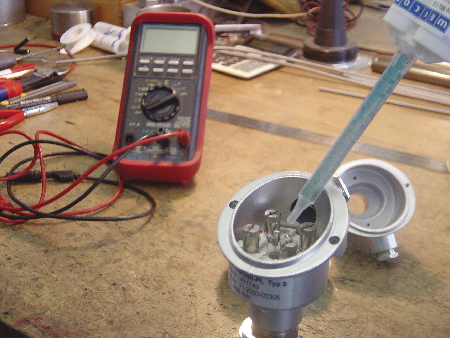 A hand is filling an electrical component with silicone on a workbench, next to a switched-on multimeter and tools.