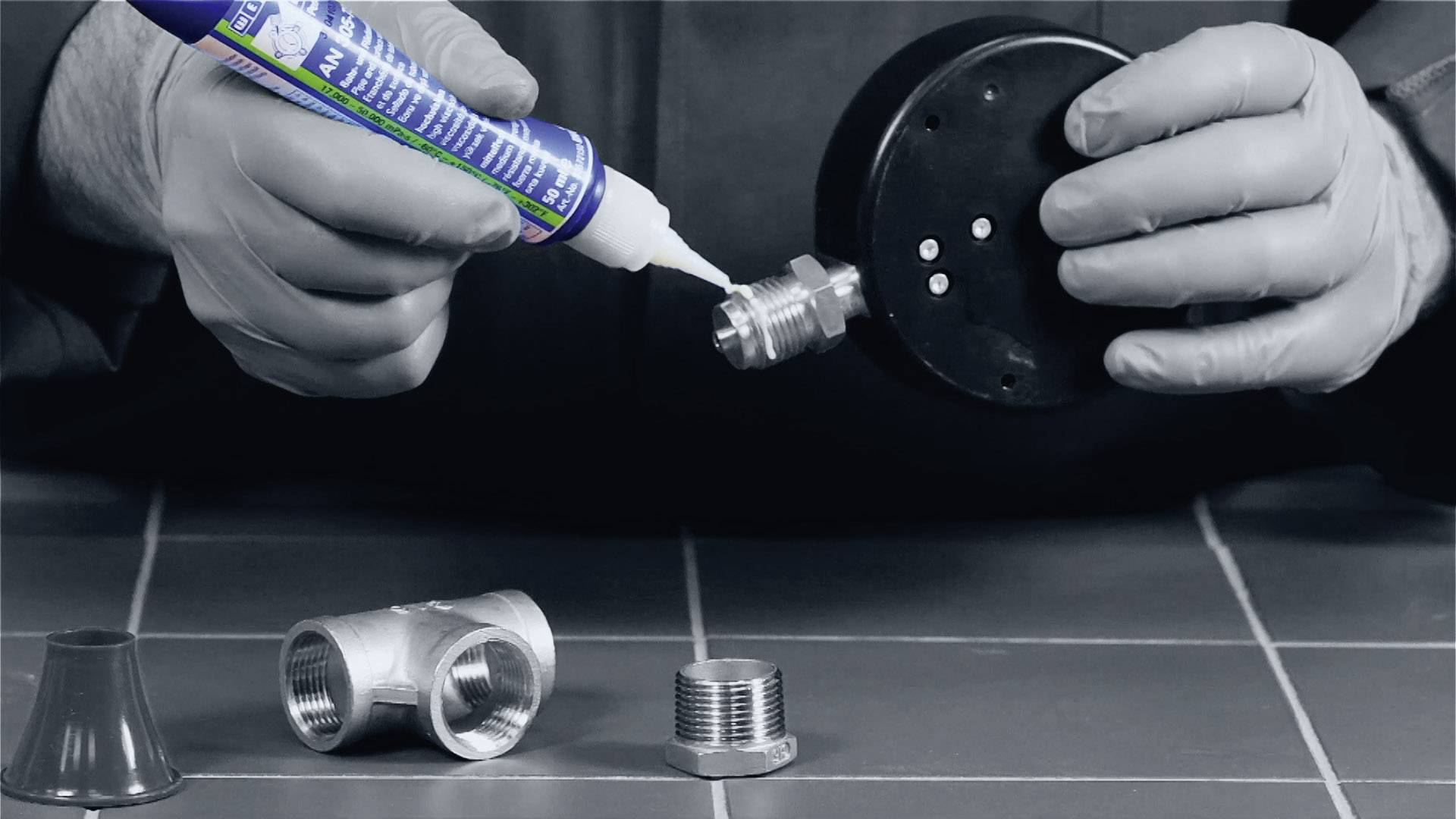A person is applying sealant to a metal thread. In the foreground, further metal components are lying on a table.
