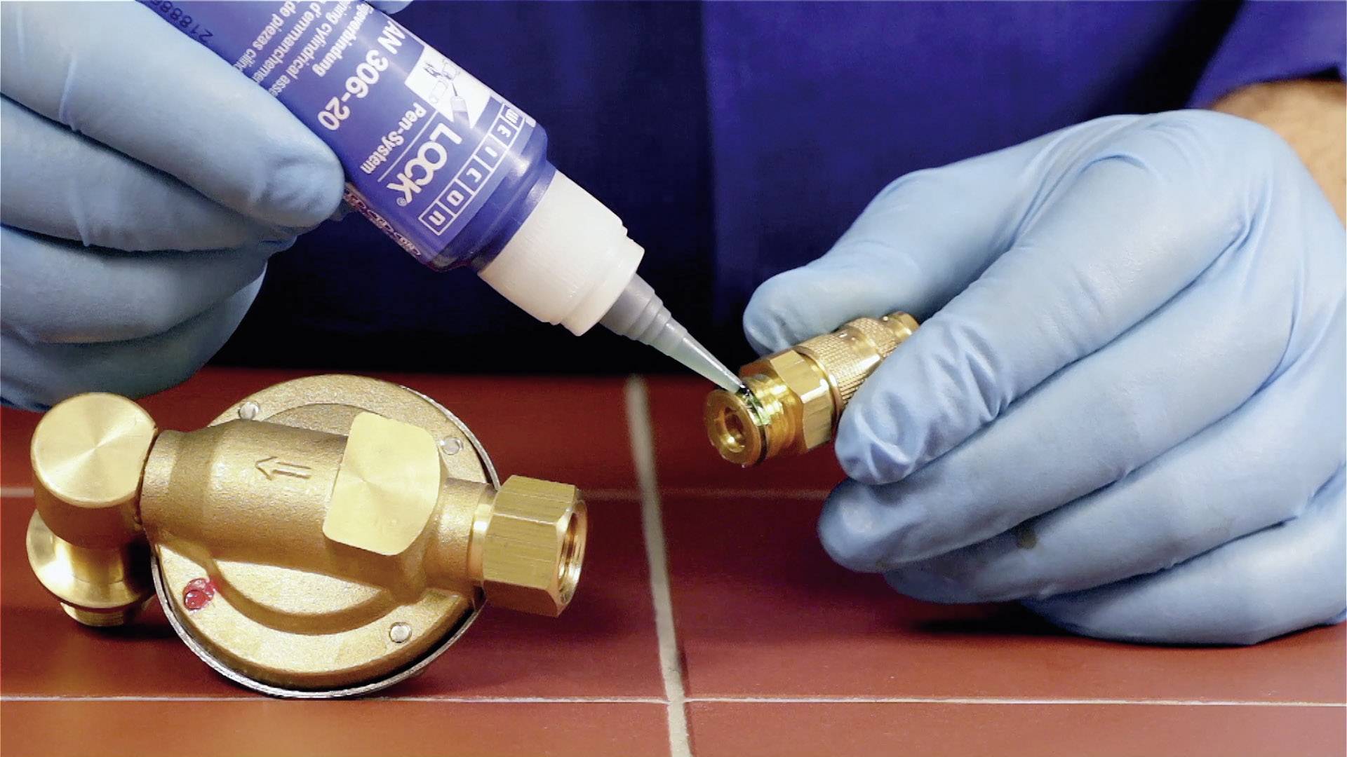 A person wearing blue gloves is applying adhesive to a small metal connector next to a valve.
