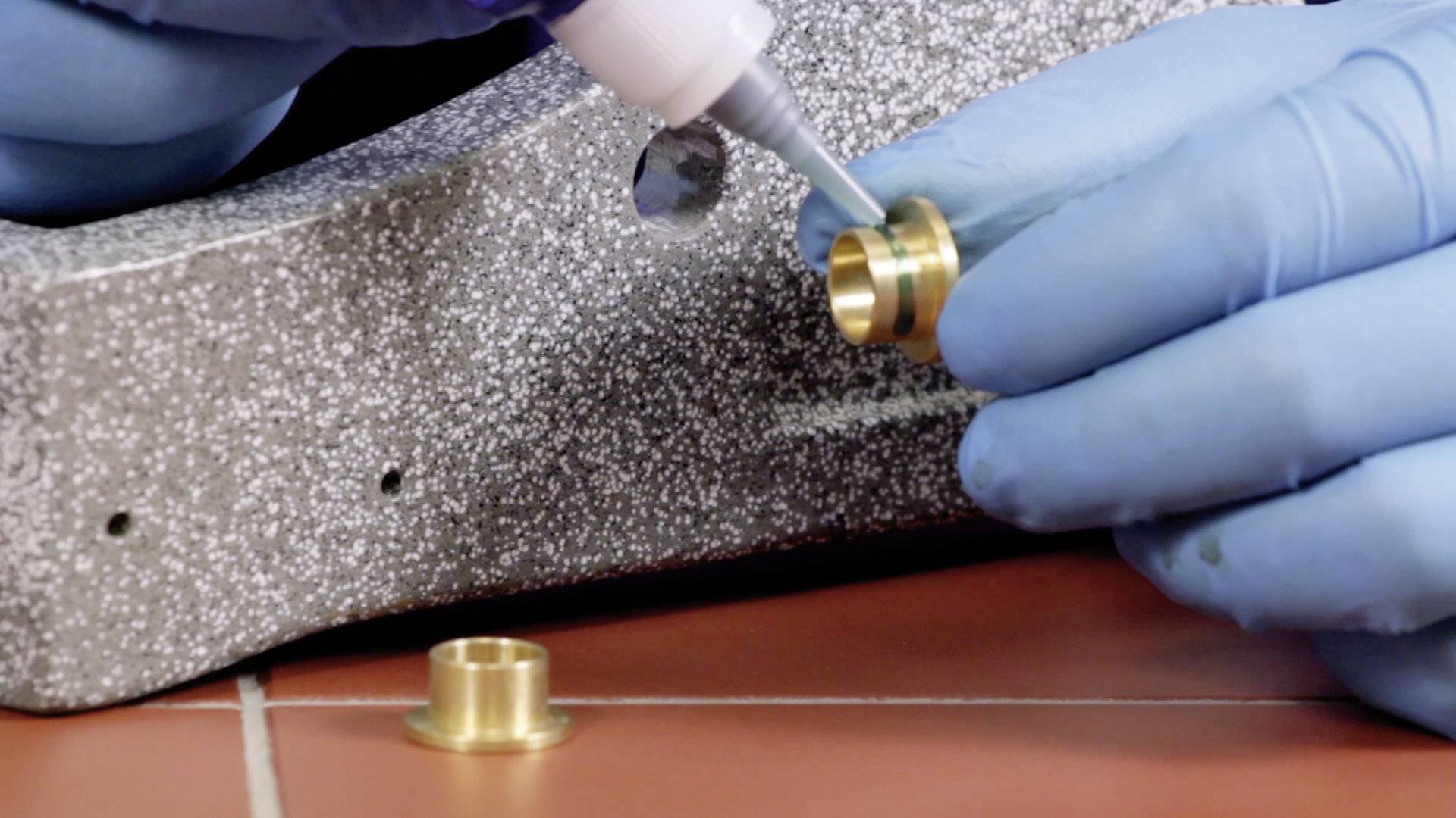 A person wearing blue gloves is applying glue to a small, metallic component. A textured surface is visible in the background.