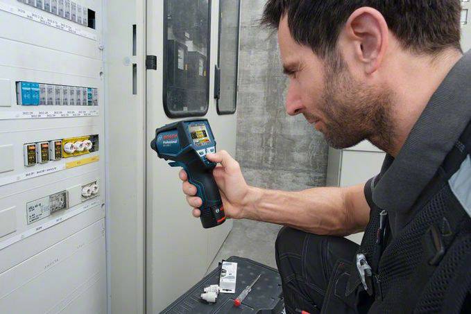A man is checking the temperature of a fuse box in a technical room using an infrared thermometer.