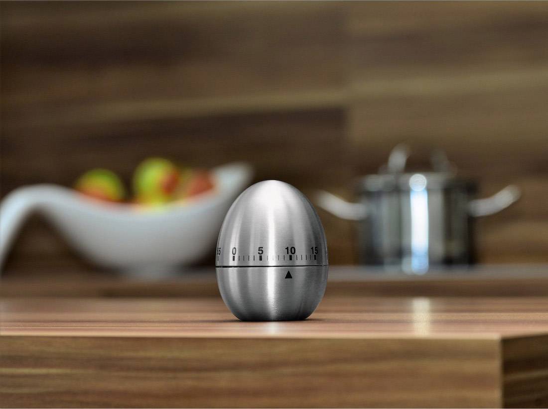 A silver egg timer in the shape of an egg sits on a wooden table. In the blurred background, a bowl of apples and a cooking pot are visible.