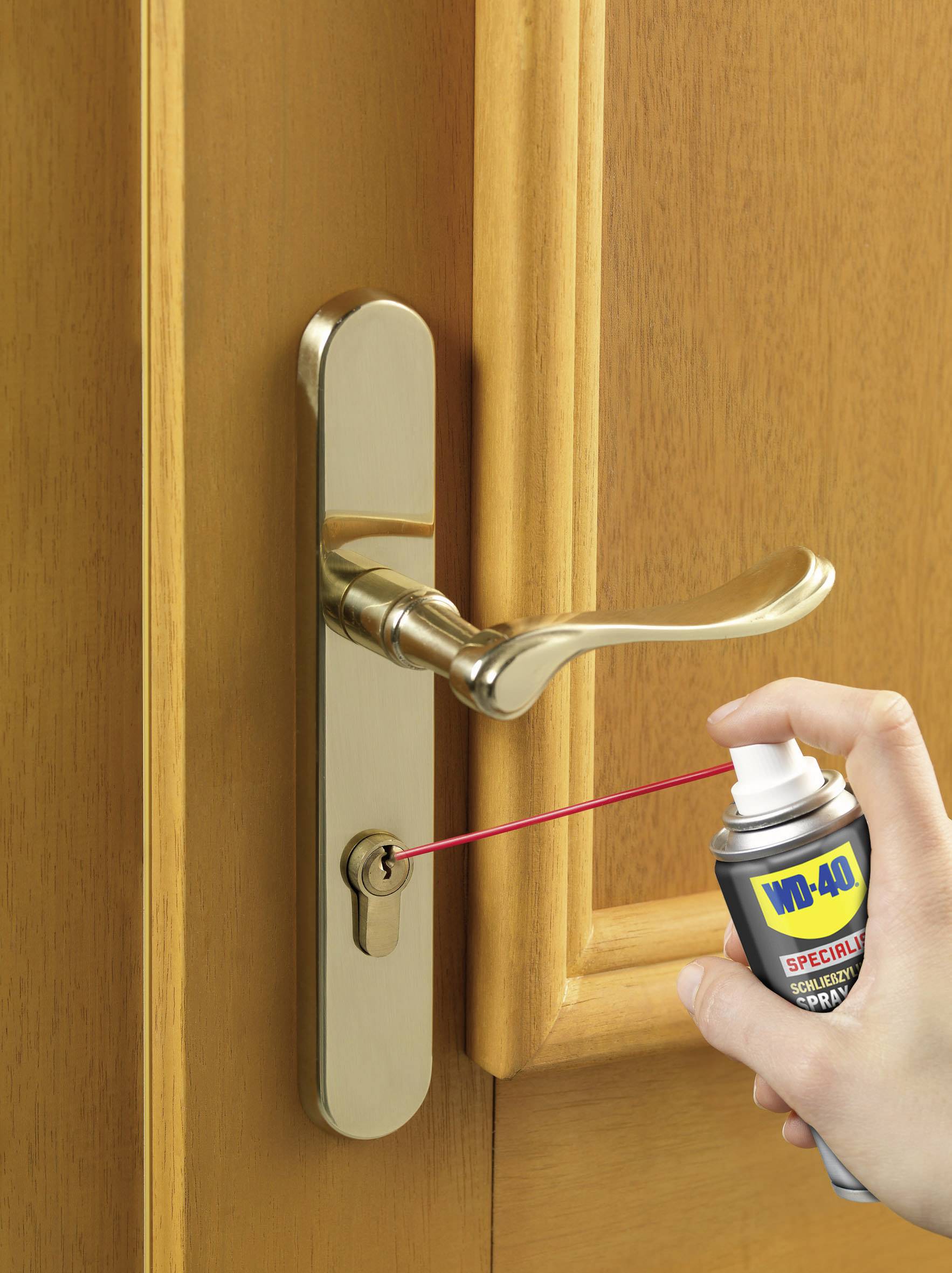 A hand sprays oil into the keyhole of a door with a golden door handle.