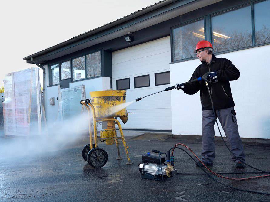 A person wearing a safety helmet is cleaning a concrete mixer with a high-pressure washer outside a warehouse.
