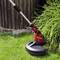 A red strimmer trims the grass at the edge of a garden in front of a wooden fence.